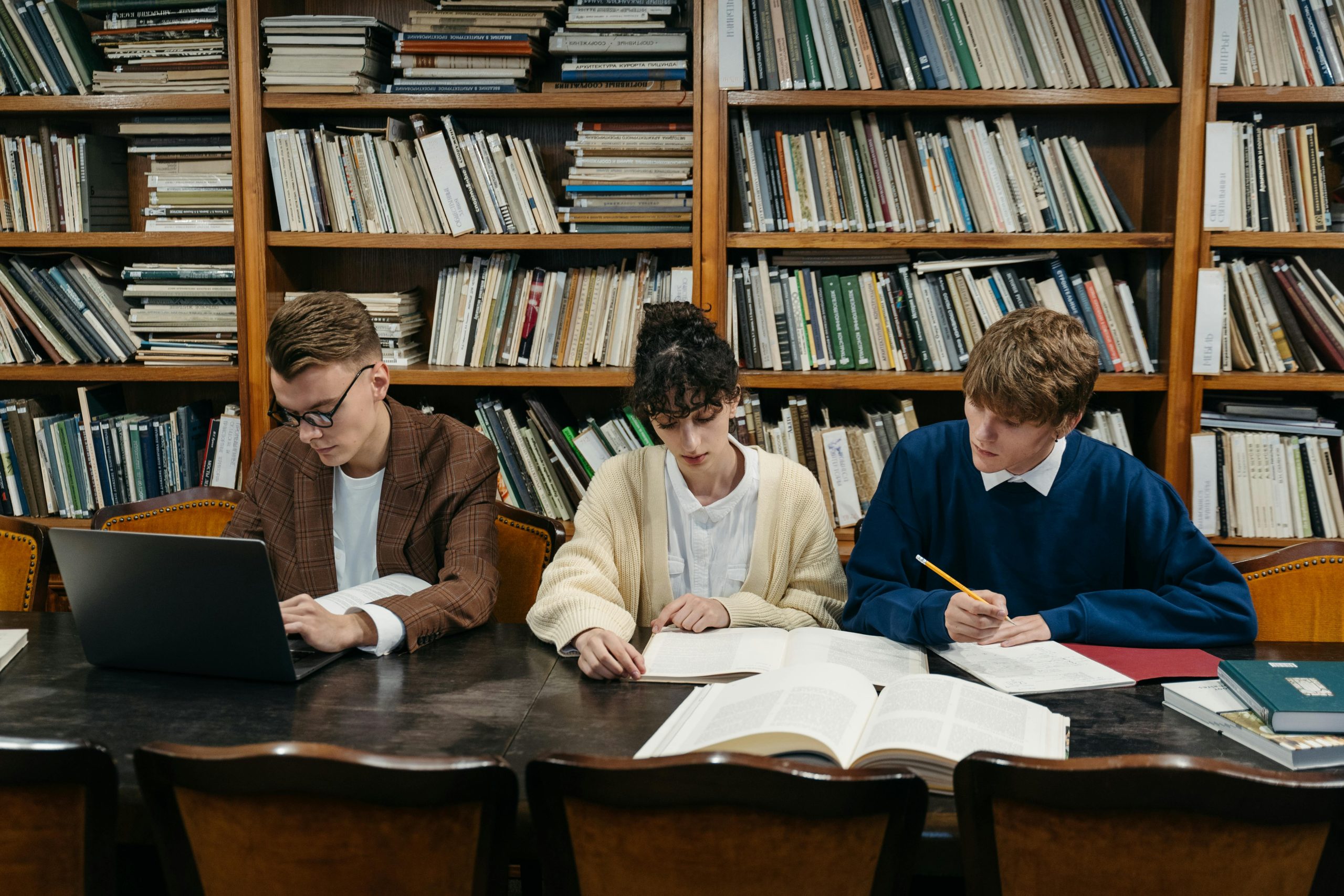 Three students work on laptops and books in a university library filled with shelves.